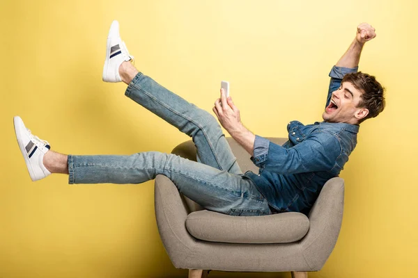 Homme heureux en utilisant smartphone assis dans le fauteuil et montrant geste gagnant sur fond jaune — Photo de stock