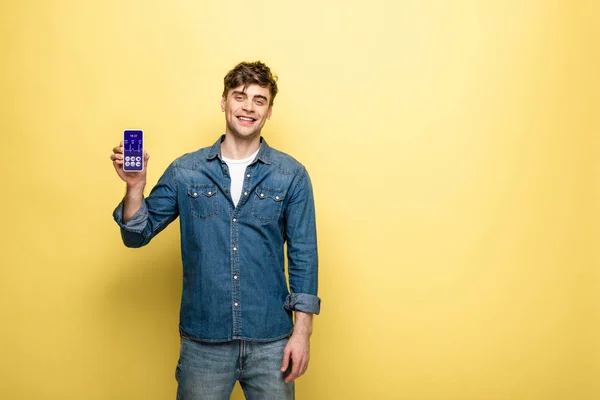 Bonito sorrindo homem em roupas jeans segurando smartphone com aplicativo de saúde no amarelo — Fotografia de Stock