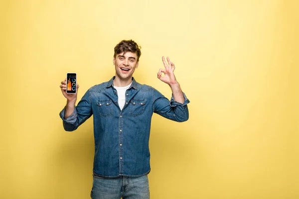 Hombre sonriente guapo en ropa de mezclilla sosteniendo teléfono inteligente con infografía y mostrando signo ok, en amarillo - foto de stock
