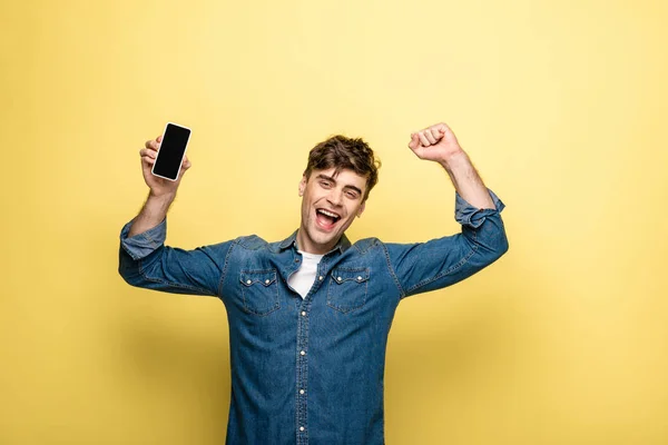 Cheerful young man holding smartphone with blank screen and showing winner gesture on yellow background — Stock Photo