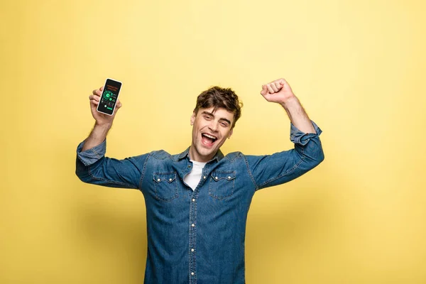 Красивый улыбающийся мужчина в джинсовой одежде держит смартфон с приложением маркетингового анализа на желтом — стоковое фото
