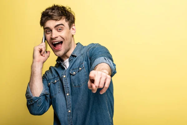 Excited young man pointing with finger at camera while talking on smartphone on yellow background — Stock Photo