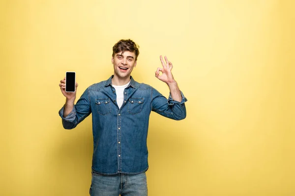 Handsome young man showing smartphone with blank screen and ok gesture while smiling at camera on yellow background — Stock Photo