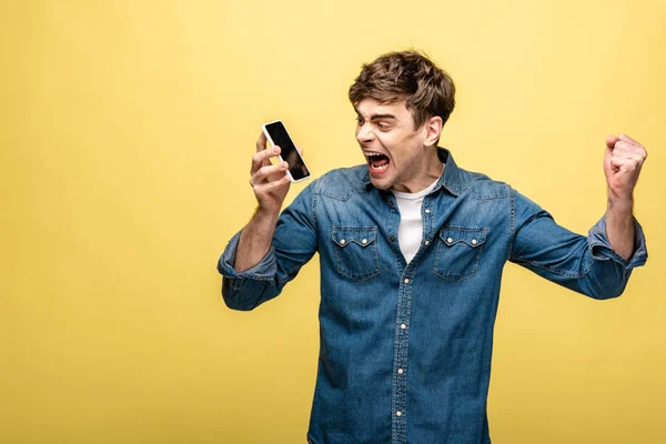 Joven irritado peleando mientras sostiene el teléfono inteligente y muestra el puño sobre el fondo amarillo - foto de stock