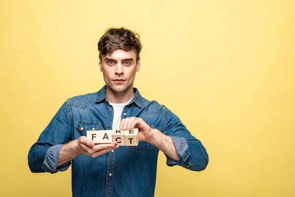 Serio joven sosteniendo cubos de madera con letras de hechos falsos sobre fondo amarillo - foto de stock