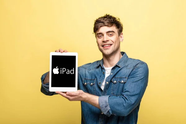 KYIV, UCRAINA - 16 MAGGIO 2019: bell'uomo sorridente in jeans che mostra tablet digitale con app iPad, isolato su giallo — Foto stock