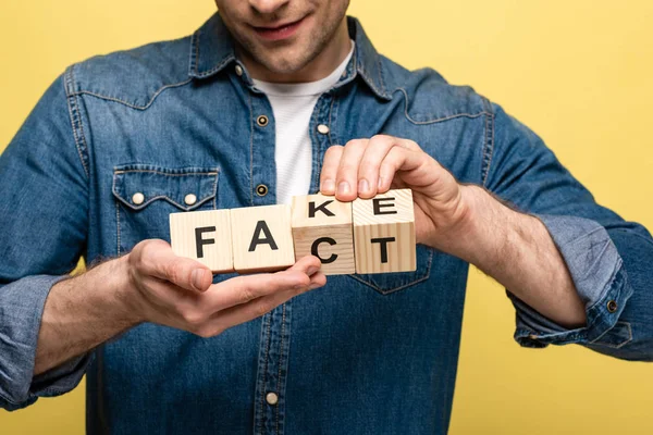 Vista cortada de homem sorridente segurando cubos de madeira com letras fato falso isolado no amarelo — Fotografia de Stock