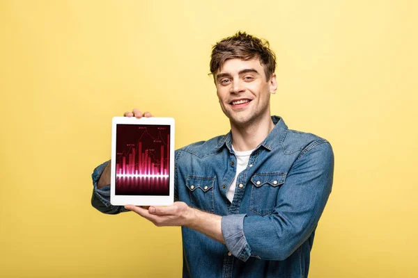 Schöner lächelnder Mann in Jeanskleidung, der ein digitales Tablet mit Grafik zeigt, isoliert auf gelb — Stockfoto