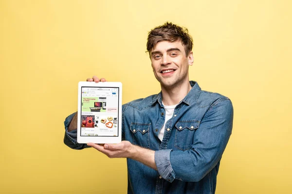 KYIV, UKRAINE - MAY 16, 2019: handsome smiling man in jeans clothes showing digital tablet with ebay app, isolated on yellow — Stock Photo