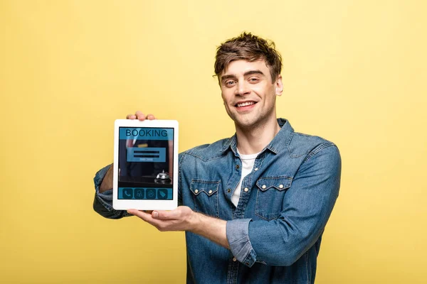 Schöner lächelnder Mann in Jeanskleidung, der ein digitales Tablet mit Buchungs-App zeigt, isoliert auf gelb — Stockfoto