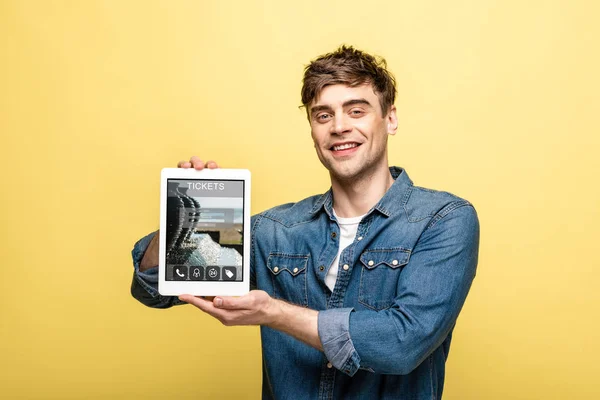 Красивый улыбающийся мужчина в джинсовой одежде показывает цифровой планшет с приложением для билетов, изолированный на желтый — стоковое фото