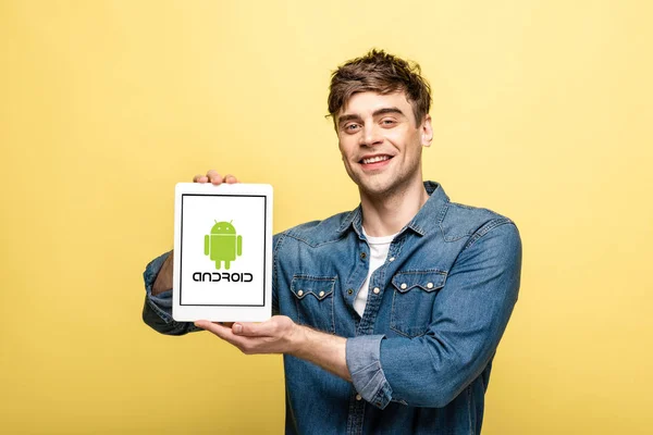 KYIV, UKRAINE - MAY 16, 2019: handsome cheerful man in jeans clothes showing digital tablet with android app, isolated on yellow — Stock Photo