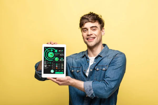 Schöner lächelnder Mann in Jeanskleidung, der ein digitales Tablet mit Infografik-App zeigt, isoliert auf gelb — Stockfoto