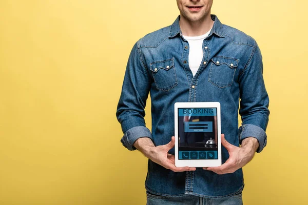 Cropped view of man in denim clothes holding digital tablet with booking app, isolated on yellow — Stock Photo