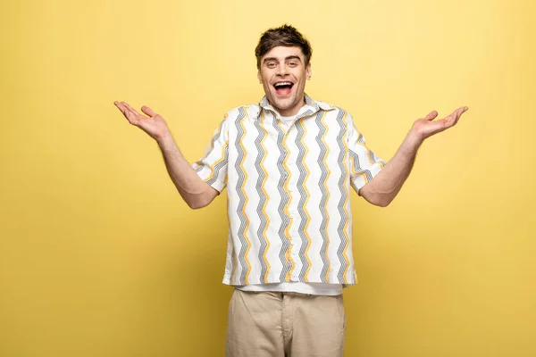 Веселый молодой человек, пожимающий плечами и улыбающийся в камеру на желтом фоне — стоковое фото