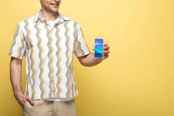 Vista recortada del hombre sonriente que muestra el teléfono inteligente con aplicación de reserva, aislado en amarillo - foto de stock