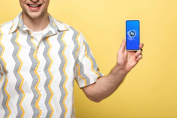 KYIV, UCRANIA - 16 de mayo de 2019: vista recortada del hombre sonriente que muestra el teléfono inteligente con la aplicación shazam, aislado en amarillo - foto de stock