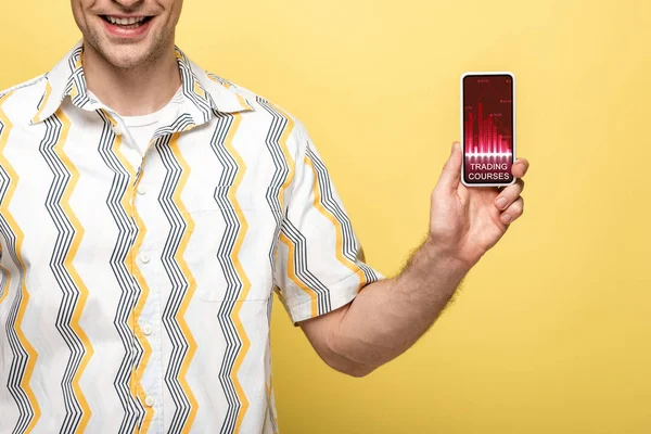 Обрезанный вид улыбающегося мужчины, показывающего смартфон с приложением для торговых курсов, изолированный на желтый — стоковое фото