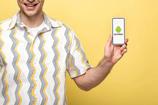 KYIV, UCRANIA - 16 de mayo de 2019: vista recortada del hombre sonriente que muestra el teléfono inteligente con aplicación para Android, aislado en amarillo - foto de stock