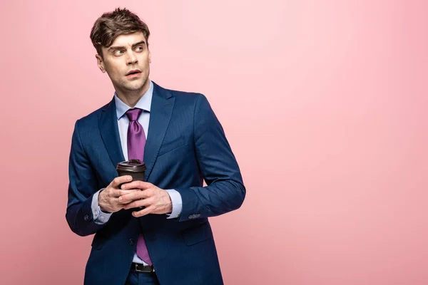 Nachdenklicher Geschäftsmann schaut weg, während er Kaffee hält, um auf rosa Hintergrund zu gehen — Stockfoto