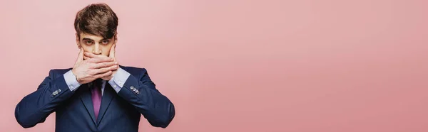 Panoramic shot of serious businessman holding hands on mouth while looking at camera  on pink background — Stock Photo