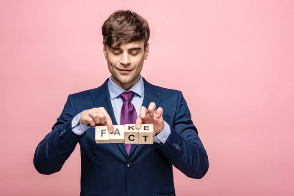 Smiling man holding wooden cubes with fake fact inscription isolated on pink — Stock Photo
