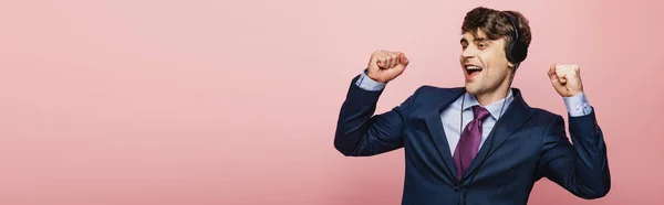Tiro panorâmico de empresário animado dançando enquanto ouve música em fones de ouvido no fundo rosa — Fotografia de Stock