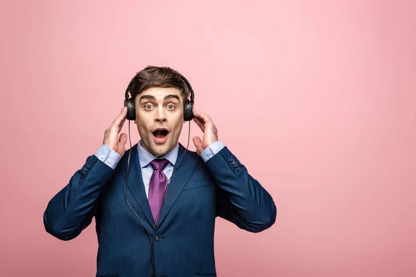 Empresário surpreso olhando para a câmera enquanto ouve música em fones de ouvido isolados em rosa — Fotografia de Stock