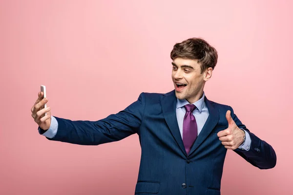 Cheerful businessman showing thumb up while taking selfie with smartphone isolated on pink — Stock Photo