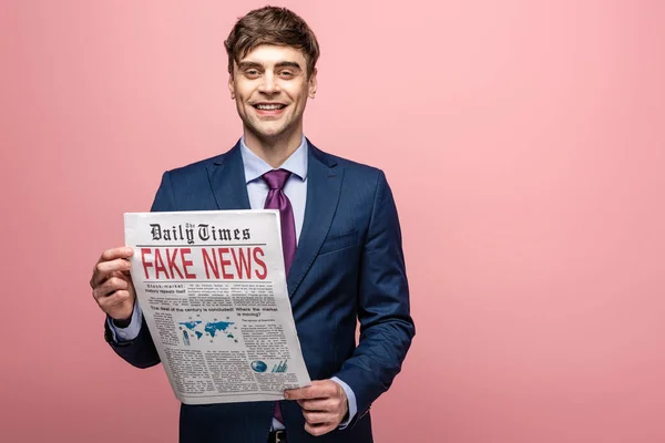 Handsome businessman showing newspaper with fake news and smiling at camera on pink background — Stock Photo