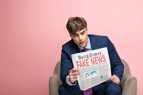 Serious businessman sitting in armchair and reading newspaper with fake news on pink background — Stock Photo