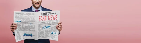 Recortado vista de sonriente hombre de negocios leyendo periódico con noticias falsas sobre fondo rosa, tiro panorámico - foto de stock