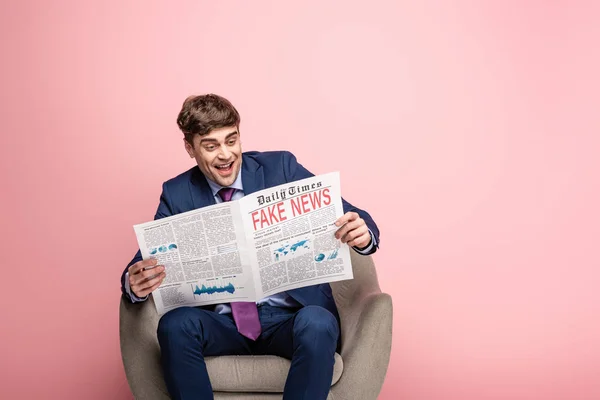 Alegre hombre de negocios sentado en sillón y leyendo el periódico con noticias falsas sobre fondo rosa - foto de stock