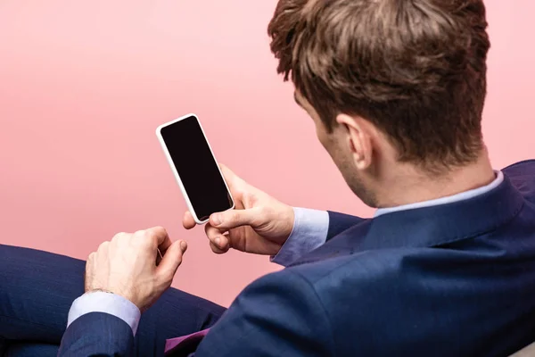 Vista trasera del hombre de negocios sentado y sosteniendo teléfono inteligente con pantalla en blanco - foto de stock