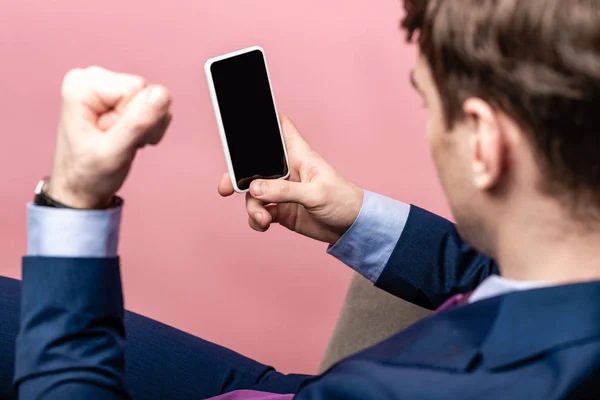 Частичный вид раздраженного бизнесмена, показывающего кулак, держа смартфон с пустым экраном, изолированным на розовом — стоковое фото