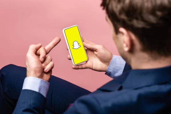 KYIV, UKRAINE - 16 de maio de 2019: visão recortada do empresário usando o smartphone com o aplicativo Snapchat e mostrando o dedo médio, isolado no rosa — Fotografia de Stock