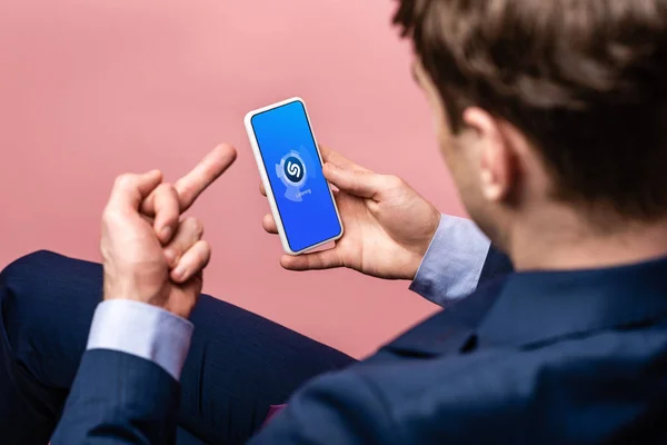 KYIV, UCRANIA - 16 DE MAYO DE 2019: vista recortada del hombre de negocios usando el teléfono inteligente con la aplicación shazam y mostrando el dedo medio, aislado en rosa - foto de stock