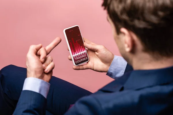 Vista recortada de hombre de negocios utilizando el teléfono inteligente con aplicación cursos de comercio mientras que muestra el dedo medio, aislado en rosa - foto de stock