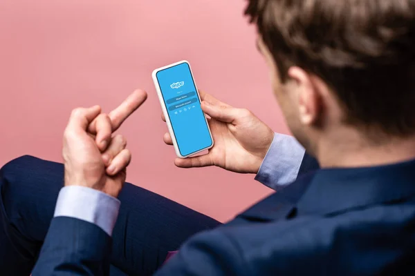 KYIV, UCRANIA - 16 DE MAYO DE 2019: vista recortada del hombre de negocios usando el teléfono inteligente con la aplicación skype y mostrando el dedo medio, aislado en rosa - foto de stock