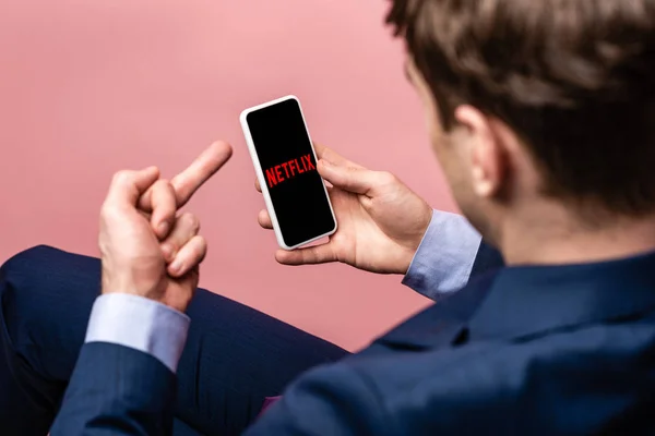 KYIV, UCRANIA - 16 de mayo de 2019: vista recortada del hombre de negocios que usa el teléfono inteligente con la aplicación netflix y muestra el dedo medio, aislado en rosa - foto de stock