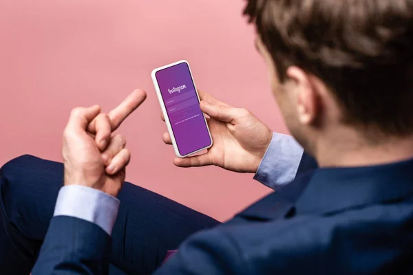 KYIV, UKRAINE - MAY 16, 2019: cropped view of businessman using smartphone with instagram app and showing middle finger, isolated on pink — Stock Photo