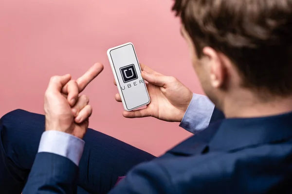 KYIV, UCRANIA - 16 DE MAYO DE 2019: vista recortada del hombre de negocios usando el teléfono inteligente con la aplicación uber y mostrando el dedo medio, aislado en rosa - foto de stock