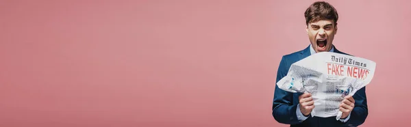 Панорамный снимок разъяренного бизнесмена, измельчающего газету фейковыми новостями, изолированными на розовом — стоковое фото