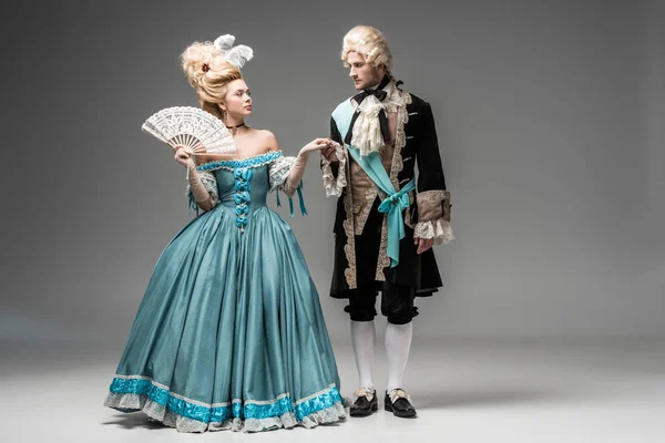 Beautiful victorian woman with fan looking at gentleman in wig while holding hands on grey — Stock Photo