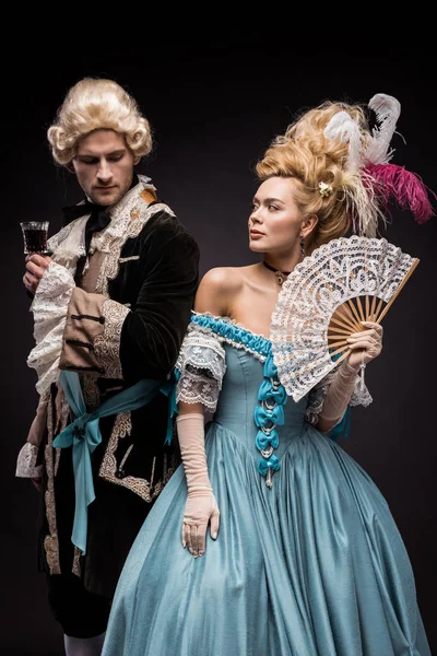 Handsome man with wine glass near young victorian woman in wig holding fan on black — Stock Photo