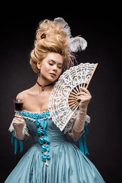 Attractive victorian woman holding fan and wine glass while standing on black — Stock Photo