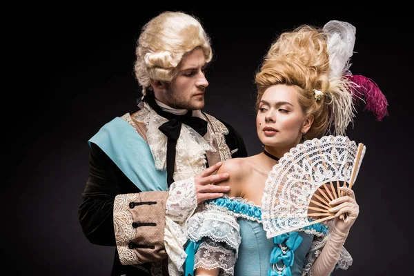 Handsome man looking at young victorian woman in wig holding fan on black — Stock Photo