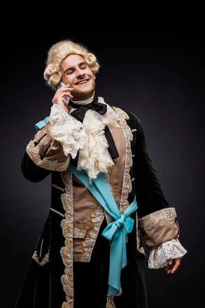 Cheerful victorian man in wig smiling while talking on smartphone on black — Stock Photo