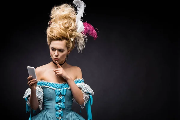 Young victorian woman in blue dress looking at smartphone and showing hush sign on black — Stock Photo
