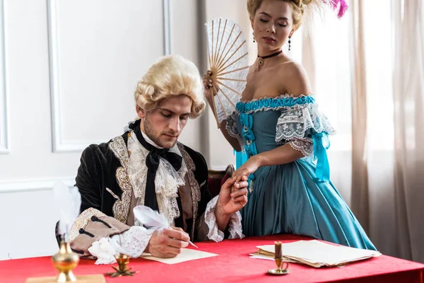 Selective focus of young victorian woman holding fan and standing near pompous man holding feather near paper — Stock Photo